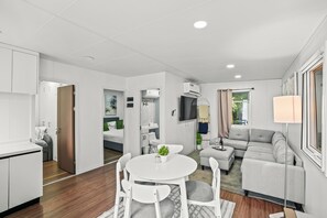 A modern, well-lit living area with a dining table, white chairs, a grey sofa, and a TV mounted on the wall. - 2BR Tiny Farm Home IN Fort Lauderdale (Hollywood)