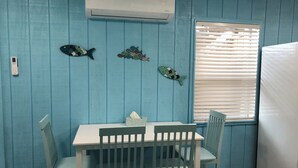 Una habitación con paredes color turquesa, una mesa blanca y sillas a juego. Hay una ventana con persianas, un equipo de aire acondicionado y decoración de peces en la pared.