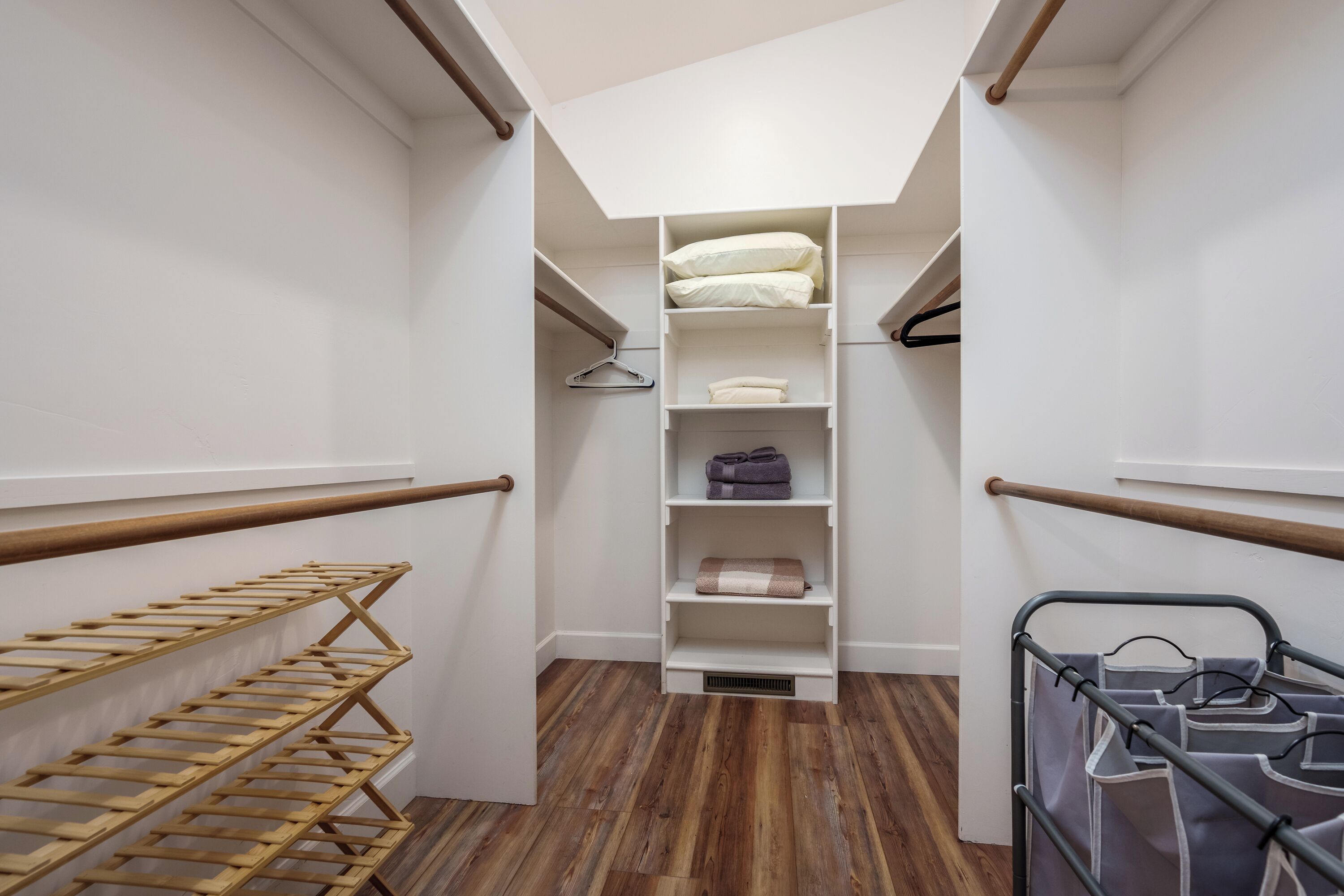 A narrow walk-in closet with wooden flooring, white walls, and a skylight. It includes shelves, a hanging rack, and a metal drying rack.