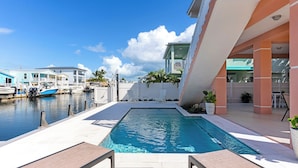 A swimming pool with lounge chairs, a marina with boats, and a modern building with a balcony. - Snowbird Paradise - New Vacation Home in Key Largo (Key Largo)
