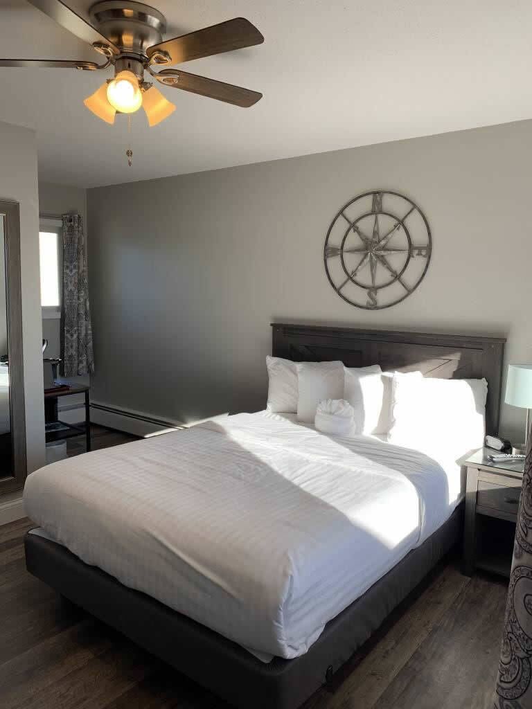 Un dormitorio con cama, ventilador de techo, reloj de pared y mesita de noche con lámpara.