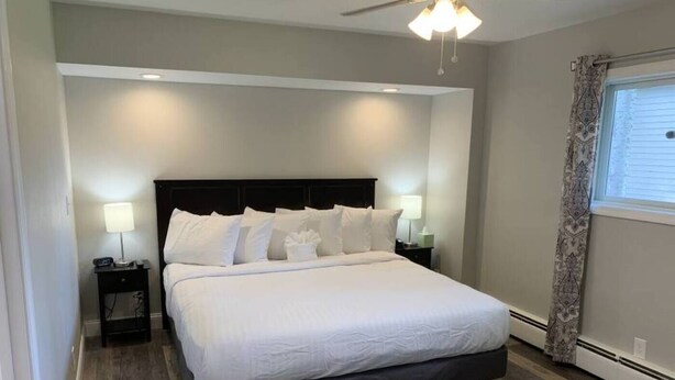 A modern bedroom with a large bed, two bedside tables with lamps, a ceiling fan, and a window with curtains. - Lakeside Motel (Ashland)