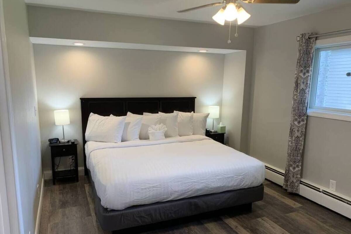 A modern bedroom with a large bed, two bedside tables with lamps, a ceiling fan, and a window with curtains.