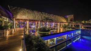 Un resort con piscina, una zona techada con techo de paja y un paseo de madera.