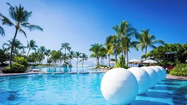 Zona de piscina con múltiples fuentes, palmeras y cielo despejado.