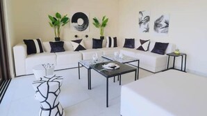 Una sala moderna con un sofá blanco, cojines blanco y negro, dos mesas auxiliares y una mesa de centro. Hay dos plantas en macetas sobre el sofá y un jarrón en la mesa de centro.