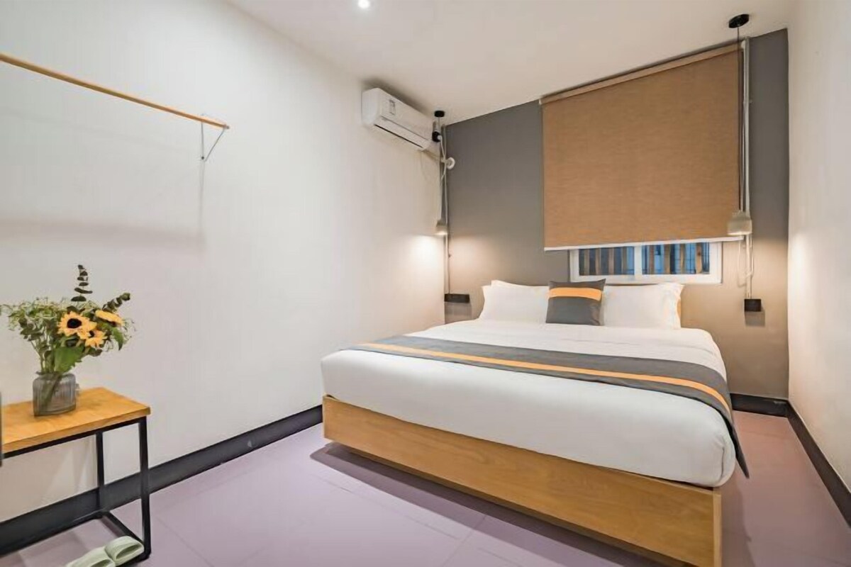 A modern hotel room with a large bed, a bedside table with a vase of flowers, a wall-mounted air conditioner, and a window with a blind.