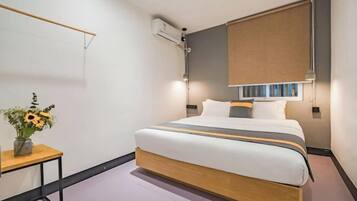 A modern hotel room with a large bed, a bedside table with a vase of flowers, a wall-mounted air conditioner, and a window with a blind.