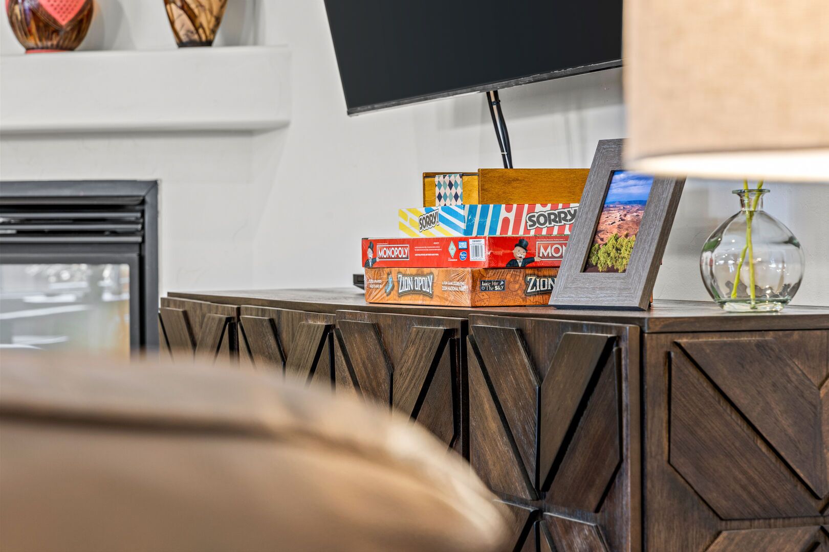 A wooden cabinet with Monopoly and ZION games, a framed picture, and a vase with a plant.