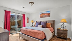 A bedroom with a bed, a nightstand, a lamp, a painting, and a sliding glass door to a balcony. - Cottonwoods 477 (Moab)