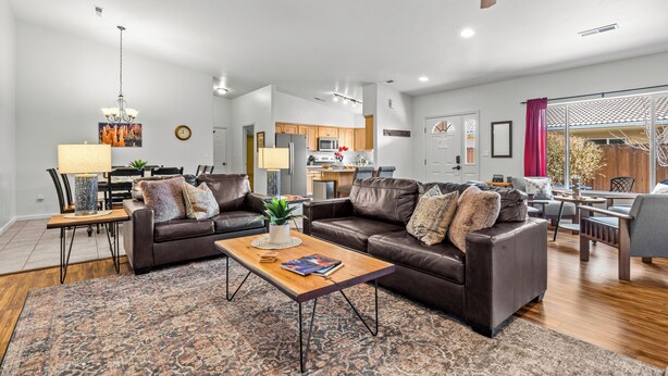 A spacious living room with a brown leather sofa, a wooden coffee table, and a patterned rug. A kitchen is visible in the background with wooden cabinets and a microwave. - Cottonwoods 477 (Moab)