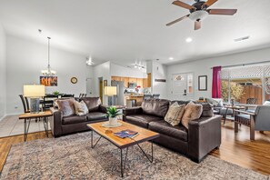 A spacious living room with a brown leather sofa, a wooden coffee table, and a patterned rug. A kitchen is visible in the background with wooden cabinets and a microwave. - Cottonwoods 477 (Moab)