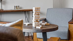 A wooden table with a stack of wooden blocks and scattered blocks on it, a grey armchair, and a wooden bench in a room with a white wall and a wooden table in the background. - Cottonwoods 477 (Moab)