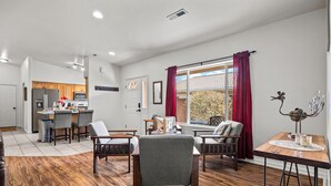 A spacious living room with a wooden floor, a dining area with a table and chairs, a kitchen with wooden cabinets, and a large window with red curtains. - Cottonwoods 477 (Moab)