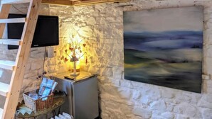 Una habitación rústica con pared de piedra, un cuadro de un paisaje, un televisor montado en la pared y una mesita con una lámpara y una cesta de objetos.