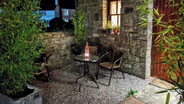 Un edificio de piedra con un pequeño patio que incluye una mesa y dos sillas, rodeado de follaje y una puerta de madera.