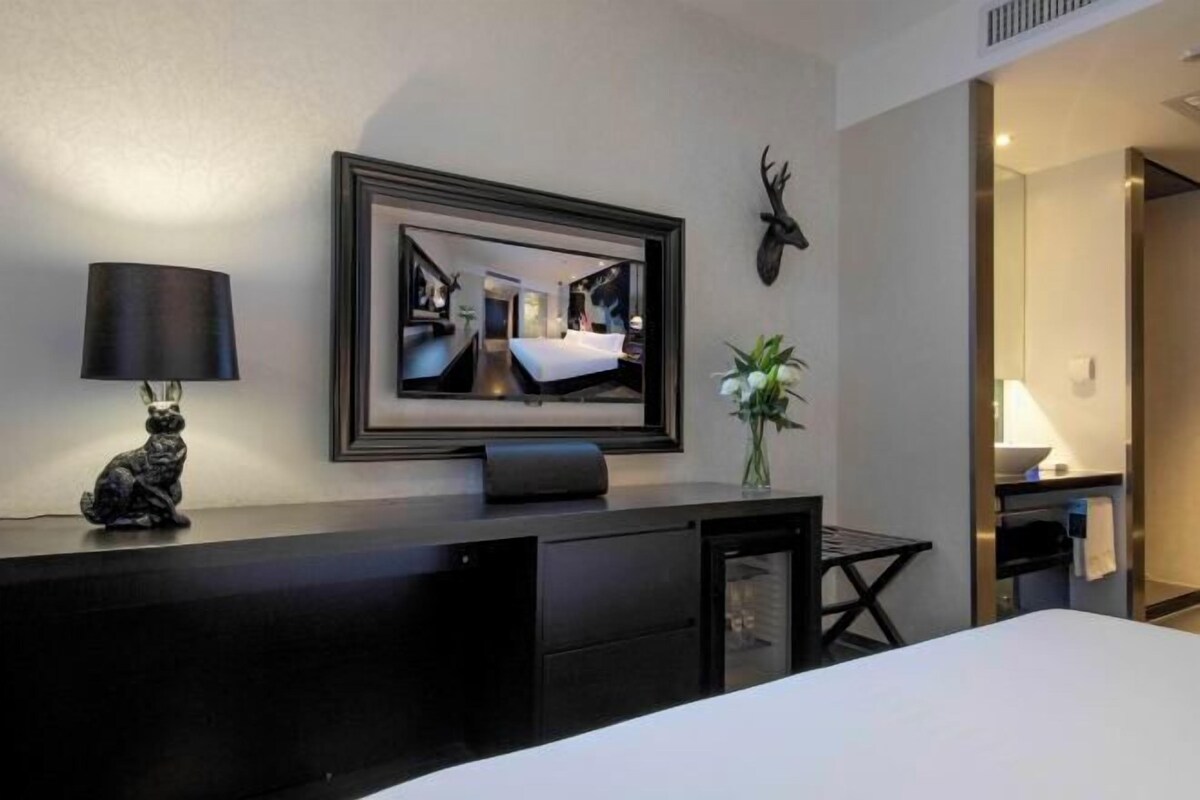 A hotel room with a bed, a desk with a lamp, a framed picture on the wall, a deer head decoration, and a vase with flowers.