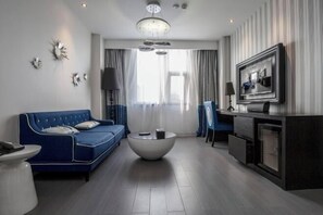 A modern living room with a blue sofa, a round coffee table, a TV mounted on the wall, and a large window with curtains. - Orange Hotel (Hefei Sanxiaokou Subway Station Store) (Hefei)