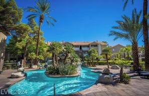 Ein Poolbereich eines Resorts mit Palmen, einer Wasserspiels und Gebäuden im Hintergrund.