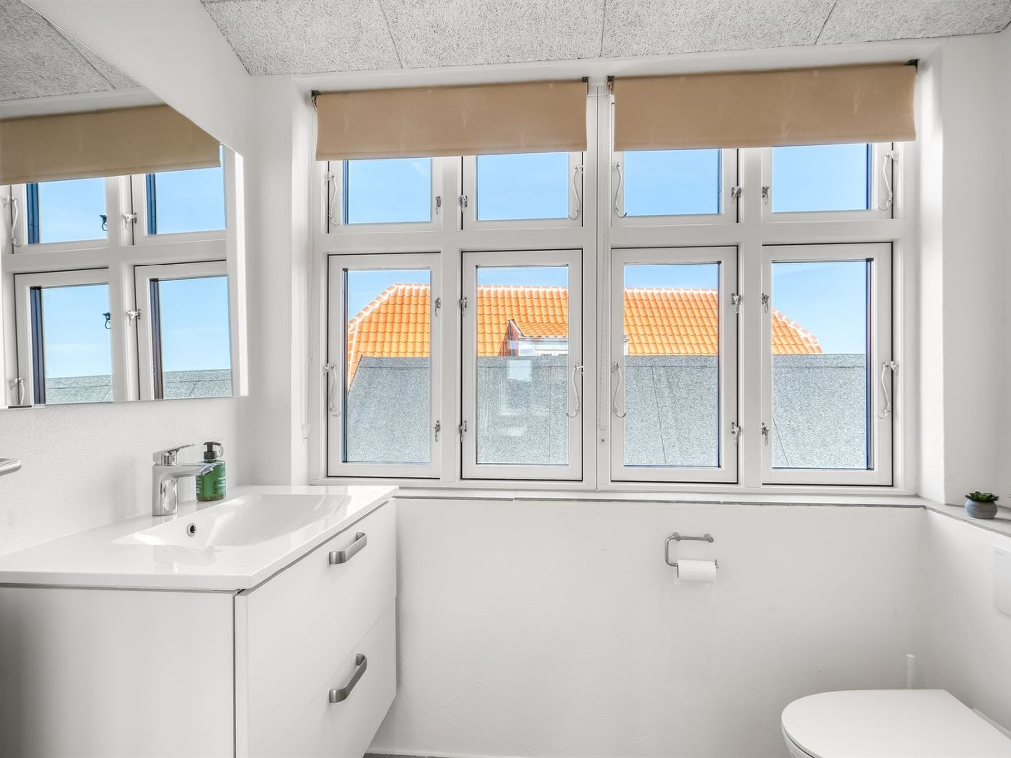 A bathroom with a white sink, two cabinets, a toilet, and a window with a view of a building with orange roofs.