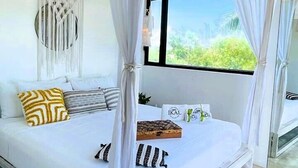 Una cama con dosel y cuatro columnas con ropa de cama y almohadas blancas, un ventilador de techo y vistas a la vegetación exterior.