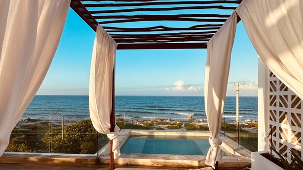 Una terraza frente a la playa con piscina, cortinas blancas y vista despejada al océano.