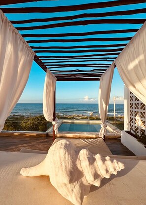 Una terraza frente a la playa con piscina, cortinas blancas y vista despejada al océano.