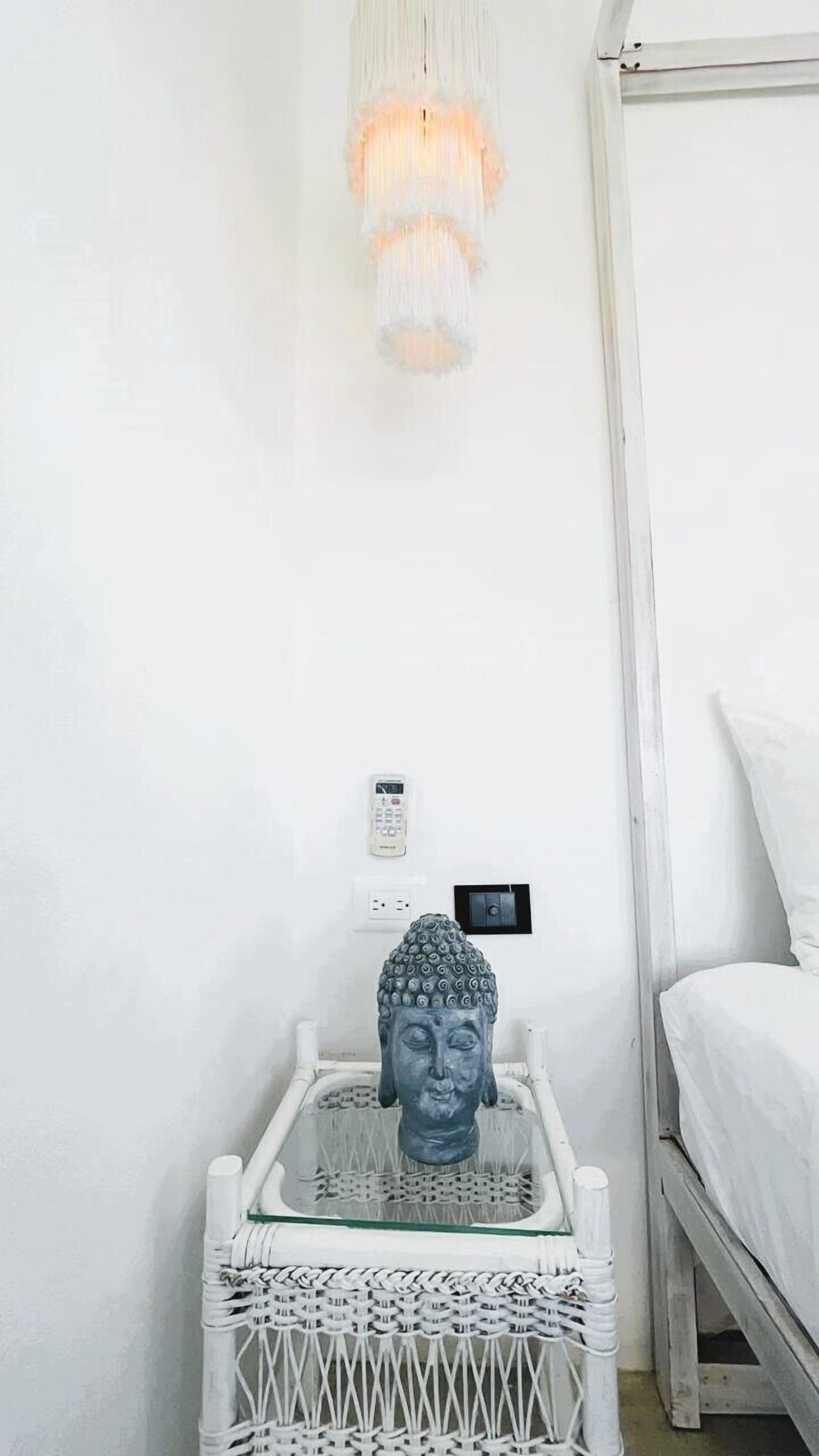 A room with a white bed, a white nightstand with a Buddha statue, a white lamp, and a power outlet on the wall.