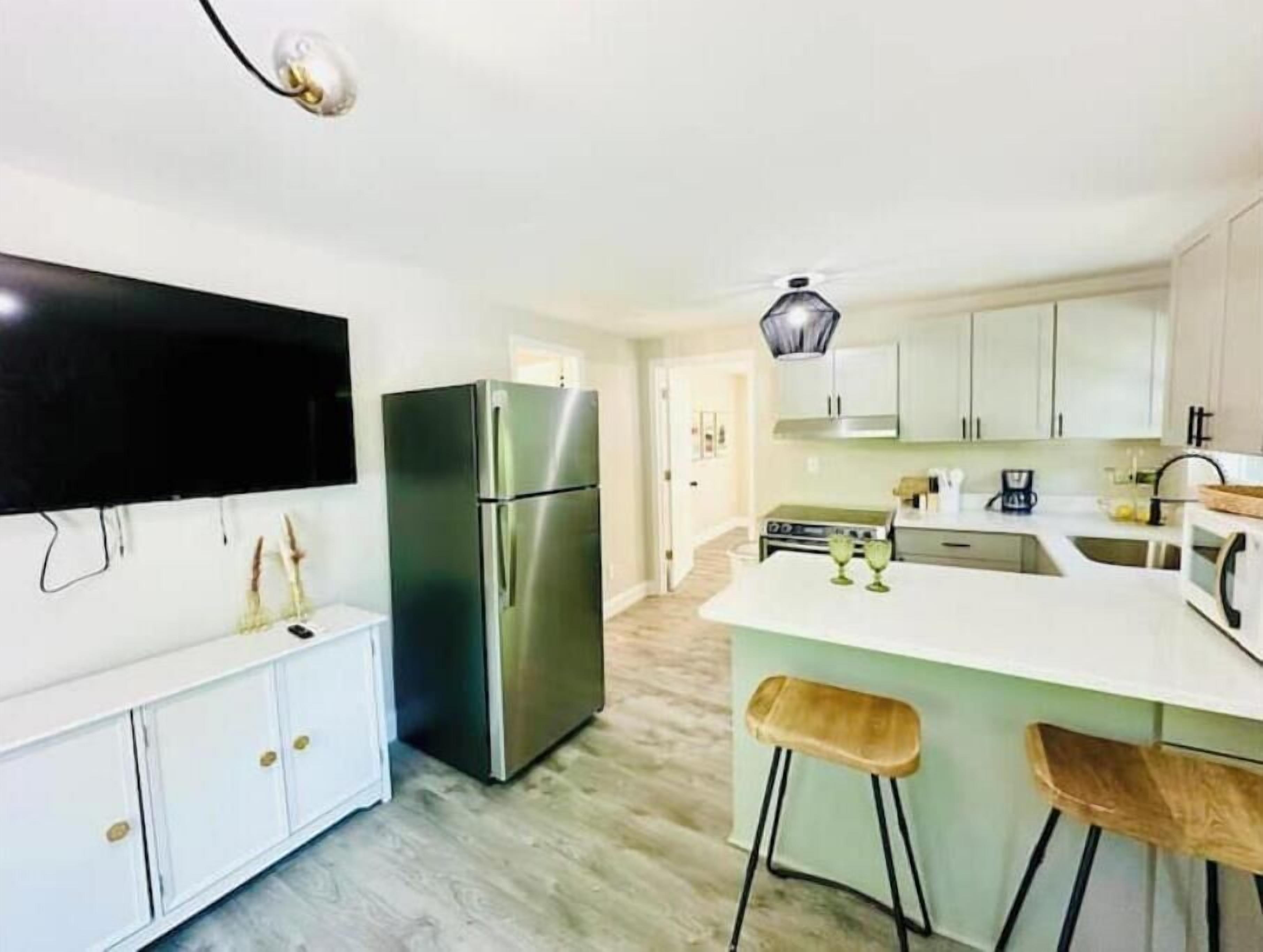 A modern kitchen with a stainless steel refrigerator, white cabinets, a large island with stools, and a flat-screen TV mounted on the wall.