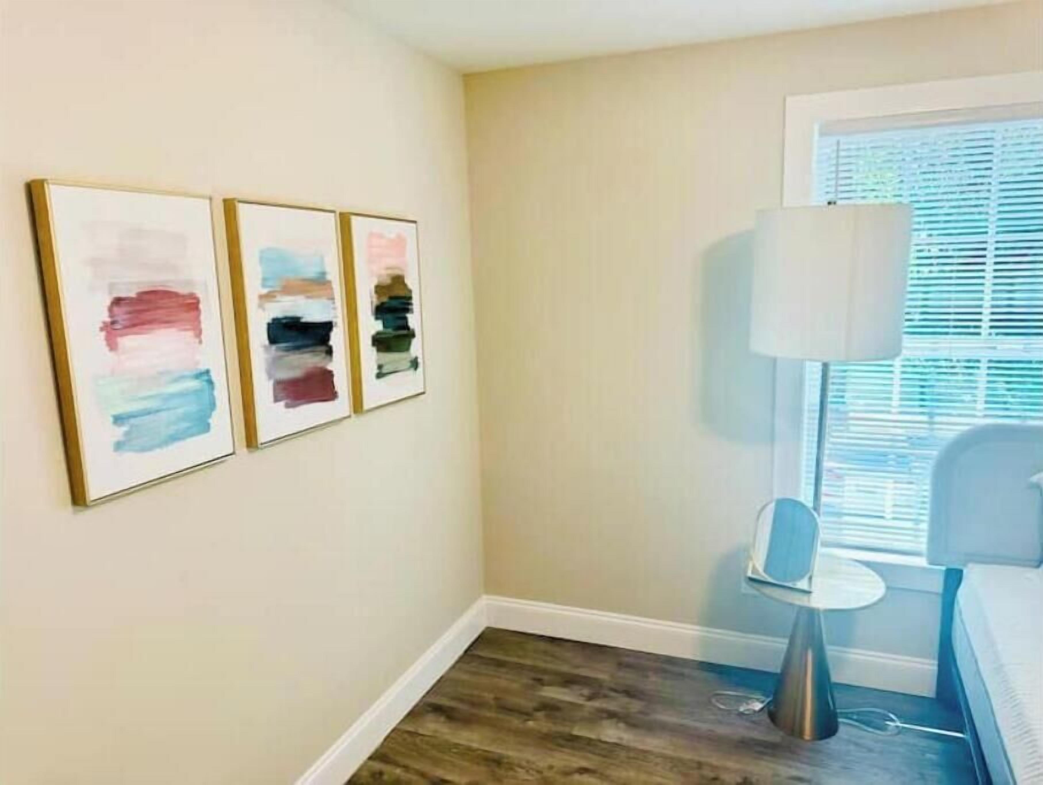 A bedroom with a bed, a bedside table, a lamp, and three abstract paintings on the wall.