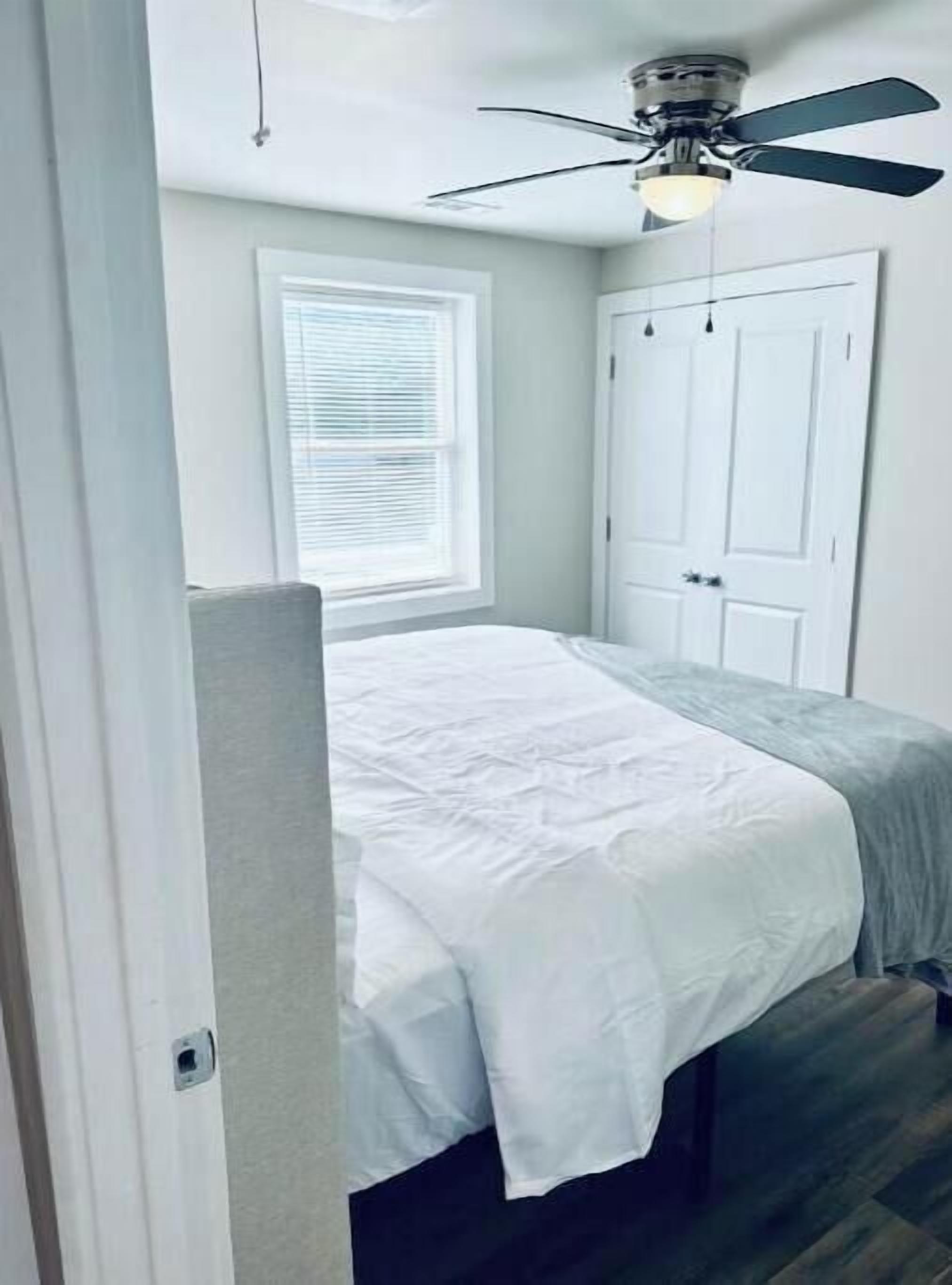 Ein Schlafzimmer mit einem Bett, einem Deckenventilator, einem Fenster mit Jalousien und einer Tür.
