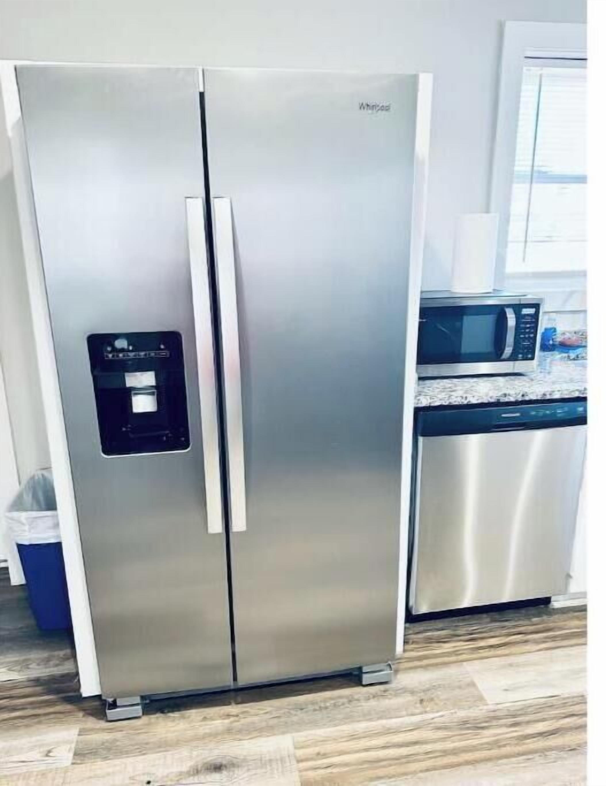 A stainless steel Whirlpool refrigerator with a water dispenser, located in a kitchen with a microwave and a trash bin.
