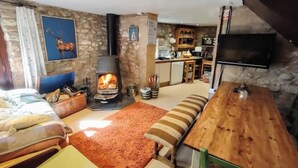 A cozy living room with a stone wall, a fireplace, a television, and a kitchen area in the background. - The Hayloft Rustic Glamping Barn, Caldbeck Village (Caldbeck)