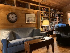 A cozy living room with a grey sofa, a wooden coffee table, a bookshelf filled with books, a wall clock, and a framed picture.