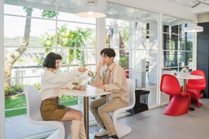 Two people seated at a table in a bright room with large windows, a red chair, and a white table.