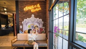 A cozy seating area with a sofa and a decorative backdrop, a large window with a view of greenery, and a neon sign above. - Inn Rose (Taoyuan)