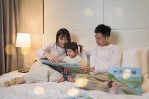 Uma família de três pessoas em uma cama, com um livro, uma luminária e uma estante fixada na parede.