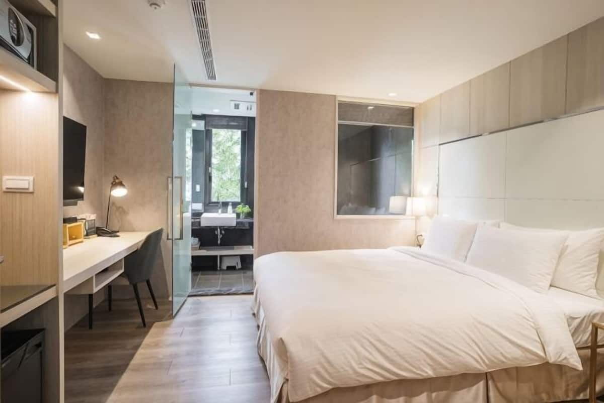 Ein modernes Hotelzimmer mit einem großen Bett, einem Schreibtisch und einem durch eine Glastür einsehbarer Badezimmer.