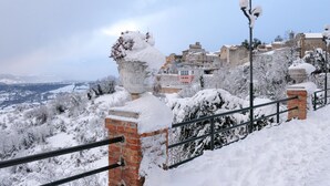 Un paisaje nevado con un muro de ladrillo y barandilla metálica, con vistas a un pueblo con edificios y árboles.