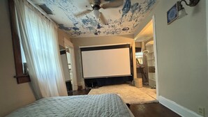 Un dormitorio con cama, ventilador de techo, pantalla de proyector y un pequeño escritorio con lámpara.