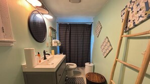Un baño pequeño con lavamanos, espejo y retrete. Un tapete estampado cubre el suelo, y una escalera de madera está apoyada contra la pared.