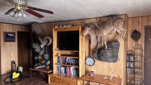 Une pièce aux murs en bois, avec un ventilateur de plafond, des têtes d’animaux empaillées accrochées au mur, une bibliothèque et un téléviseur.