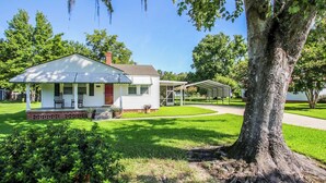 A single-story house with a porch, a covered patio, and a large tree in the foreground. - Miss Audrey’s House is a charming cottage in the heart of historic Richmond Hill (Richmond Hill)
