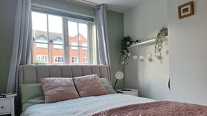 Quarto com cama, mesa de cabeceira, estante na parede com plantas e decorações, cortinas na janela e vista para edifícios.