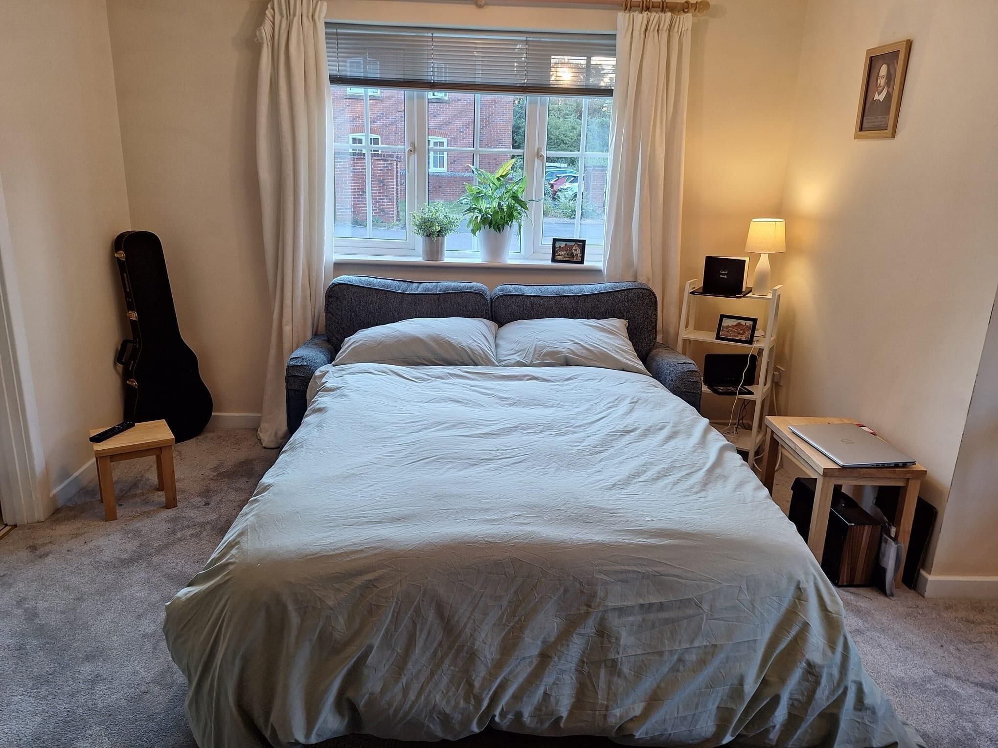 Un dormitorio con una cama, una guitarra, una lámpara y un ordenador portátil sobre una mesita.