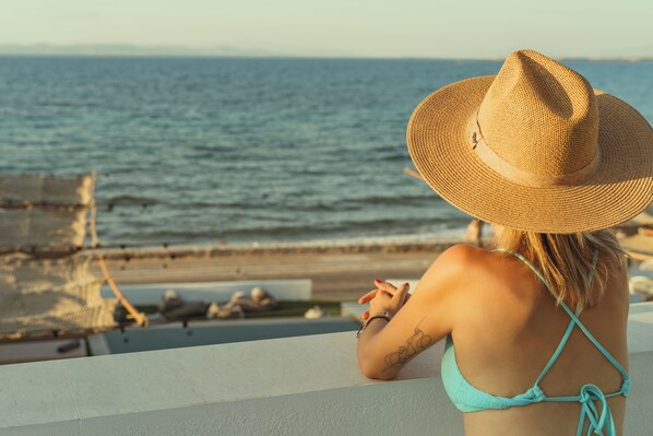 Una persona con traje de baño turquesa y sombrero de paja mirando el océano desde un balcón.