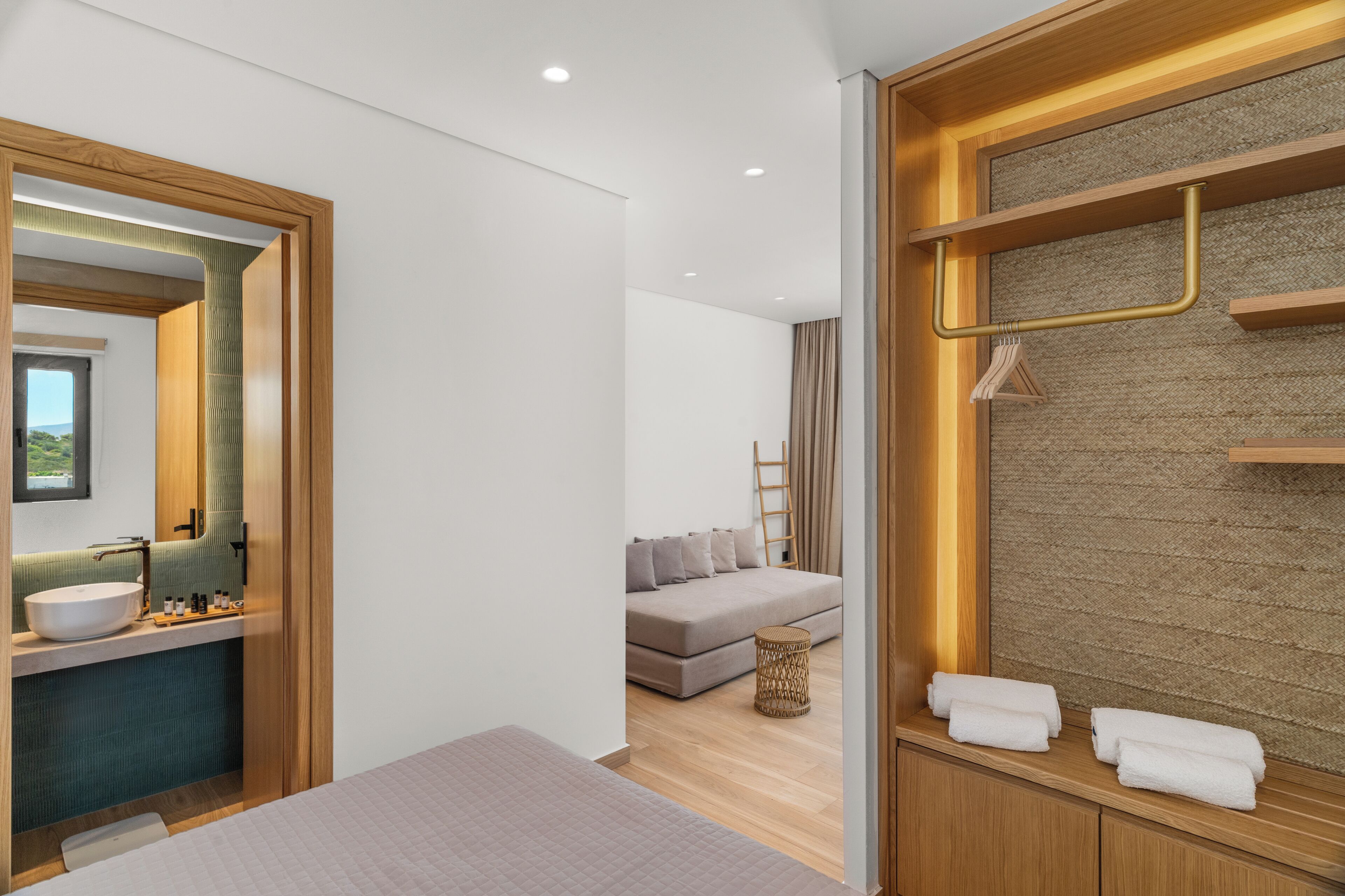 A modern hotel room with a large bed, a bathroom with a sink and mirror, a sofa, and a wall-mounted towel rack.