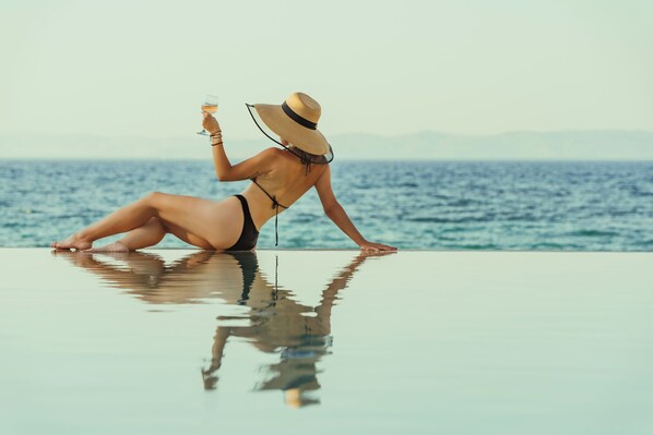 Una persona en traje de baño sentada al borde de una piscina, con una bebida, mirando un cuerpo de agua.