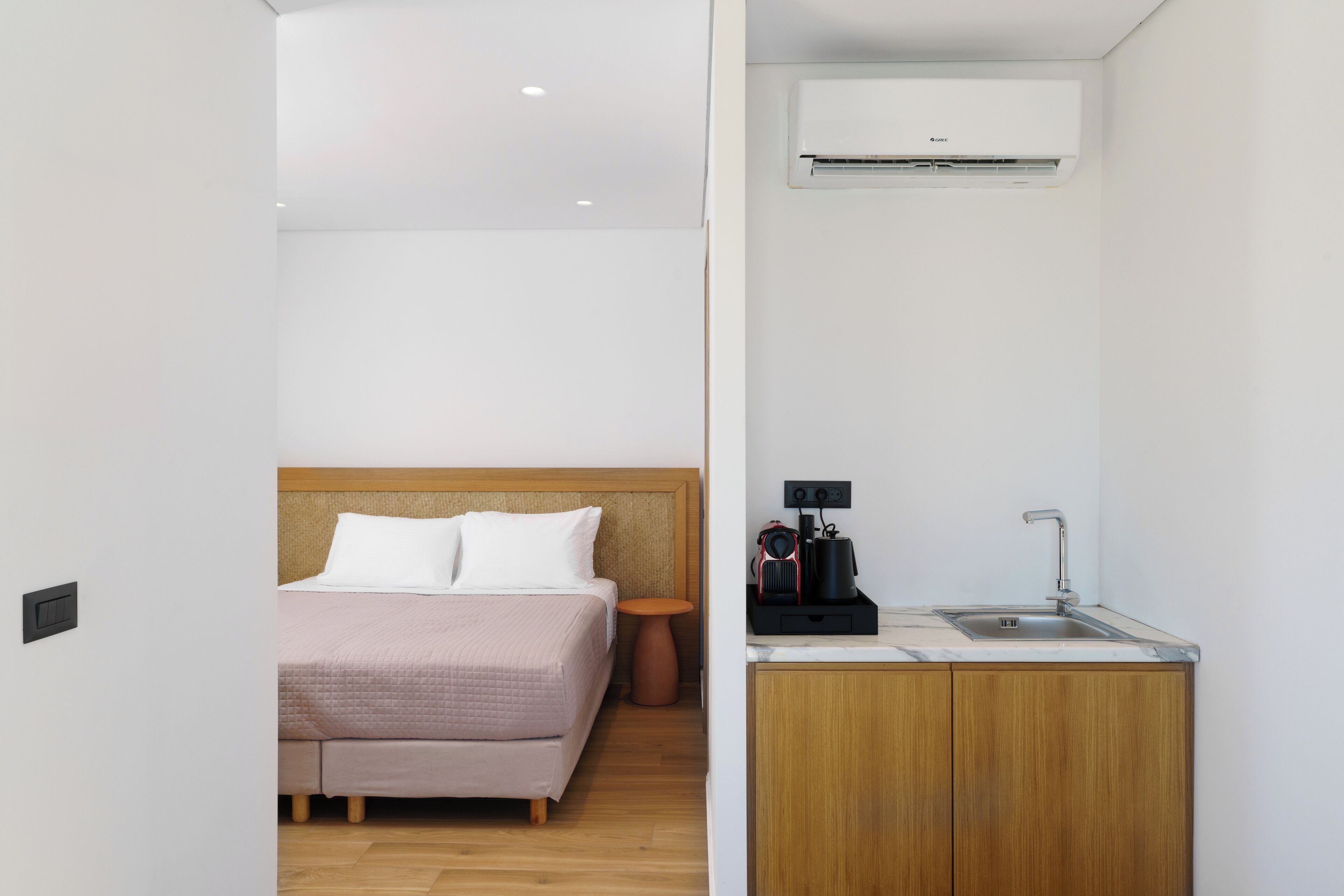 A compact hotel room with a bed, a small wooden nightstand, a single round table, a kitchenette with a sink, and an air conditioning unit mounted on the wall.
