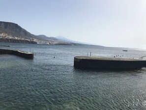 Un paysage côtier avec des eaux calmes, une jetée en béton et une chaîne de montagnes à l’horizon.